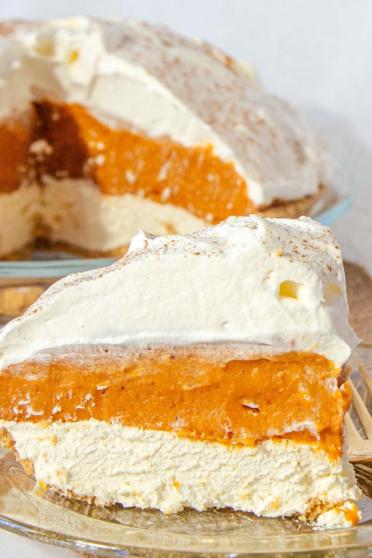 Slice of pumpkin pie with layers of pumpkin, cream cheese and whipped cream on amber coloured glass plate. Whole pie with slice out in background.