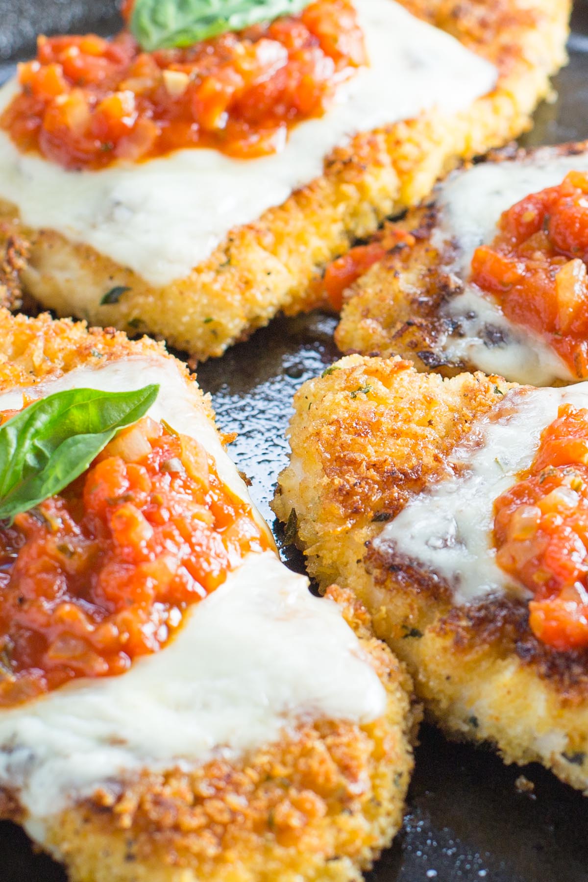 Cast iron skillet with chicken cutlets topped with marinara sauce melted cheese and fresh basil leaf.