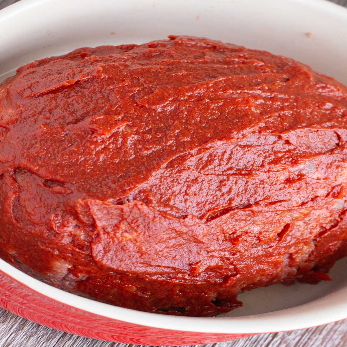 Unbaked meatloaf covered in tomato paste in oval baking dish.