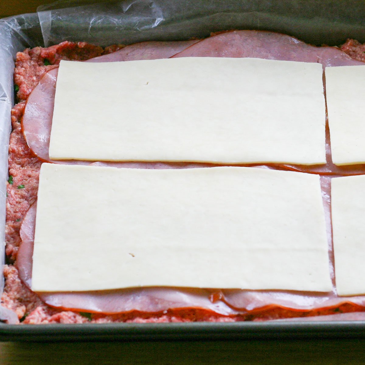 Ground beef mixture in baking sheet topped with ham and mozzarella slices.
