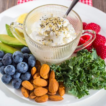 Cup of yogurt sprinkled with seeds on white plate of berries, almonds, avocado and kale.