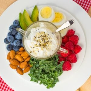 Cup of yogurt sprinkled with seeds on white plate of berries, almonds, avocado and kale.