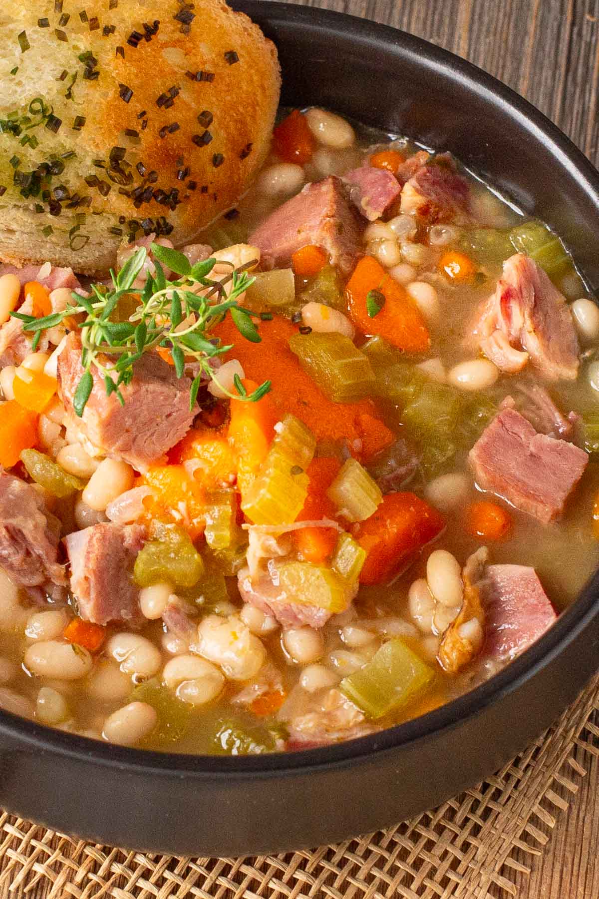 Black bowl of soup with ham, bean and vegetables and side of garlic bread.