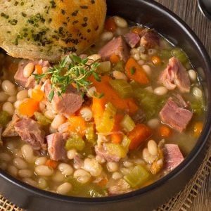 Black bowl of soup with ham, bean and vegetables and side of garlic bread.