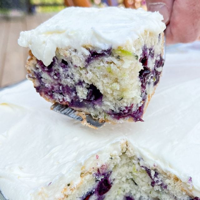 Close-up of a slice of blueberry cake with creamy white frosting being lifted from the cake pan.