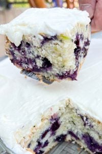 Close-up of a slice of blueberry cake with creamy white frosting being lifted from the cake pan.