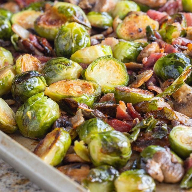 Pan of roasted Brussels sprouts with crispy edges, mixed with browned bacon bits and mushrooms.