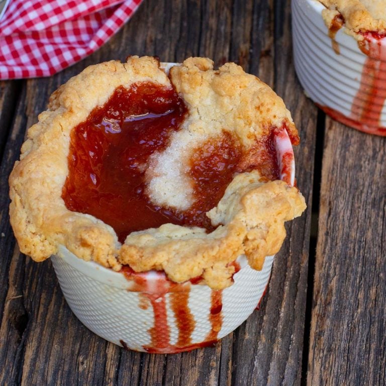 Strawberry rhubarb pie in a white ramekin with golden crust, overflows with red filling on rustic wood.