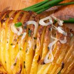 Golden Hasselback potatoes with crispy layers on a wooden board garnished with chives and sliced shallots.