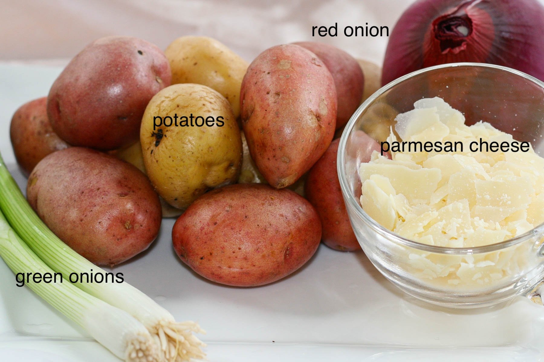 A selection of ingredients on a table: red potatoes, green onions, a whole red onion, and shaved Parmesan cheese in a glass bowl.