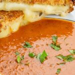 Grilled cheese sandwich with melting cheese on golden bread, paired with a bowl of vibrant tomato soup garnished with fresh parsley.