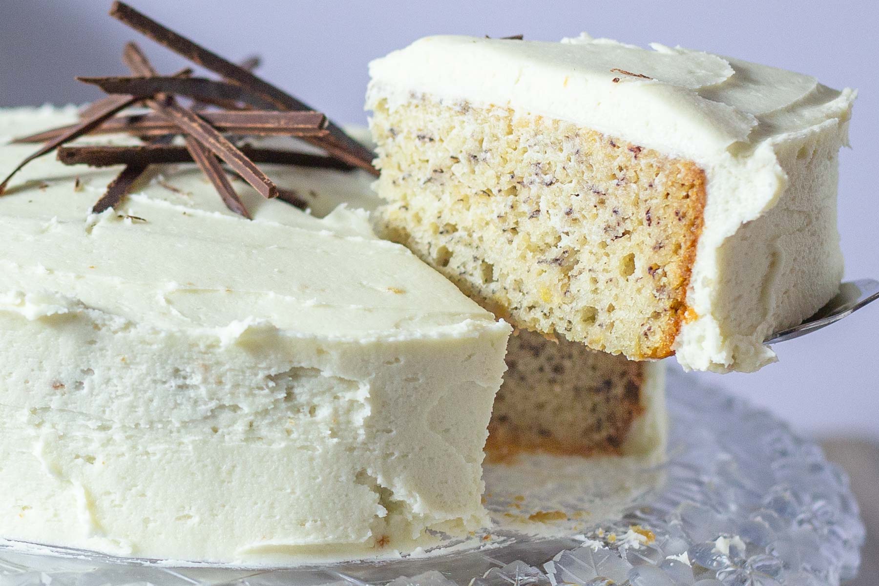 A slice of banana cake with white frosting is lifted from a cake decorated with chocolate slivers on a crystal platter.
