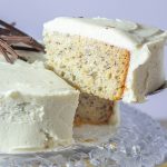 A slice of banana cake with white frosting is lifted from a cake decorated with chocolate slivers on a crystal platter.