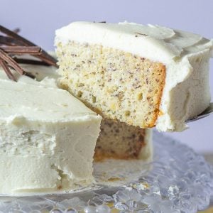 A slice of banana cake with white frosting is lifted from a cake decorated with chocolate slivers on a crystal platter.
