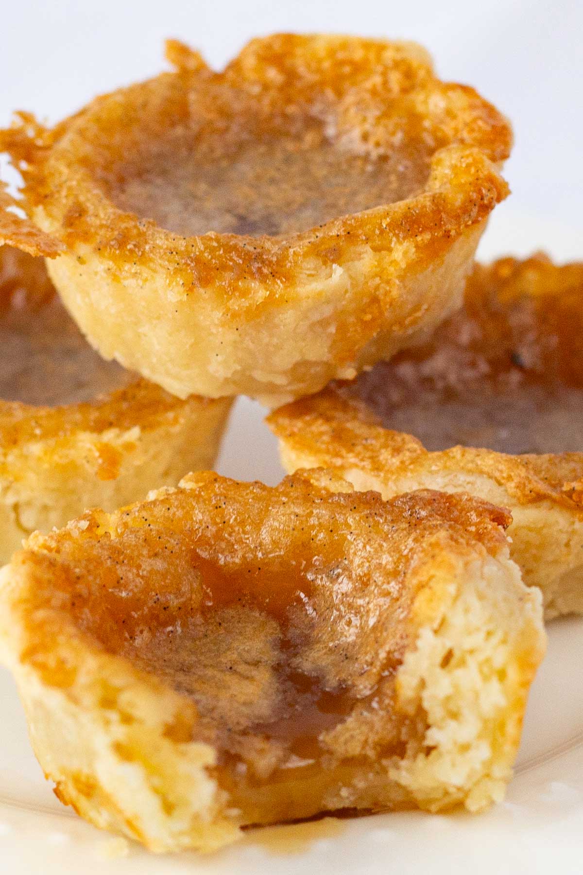 Close-up of golden-brown butter tarts with flaky crusts and gooey caramel centers, stacked on a plate.