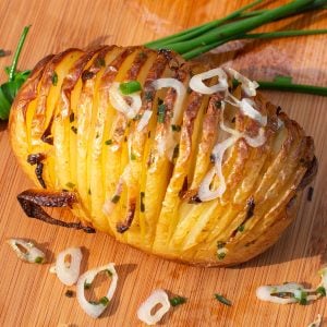 Golden Hasselback potatoes with crispy layers on a wooden board garnished with chives and sliced shallots.