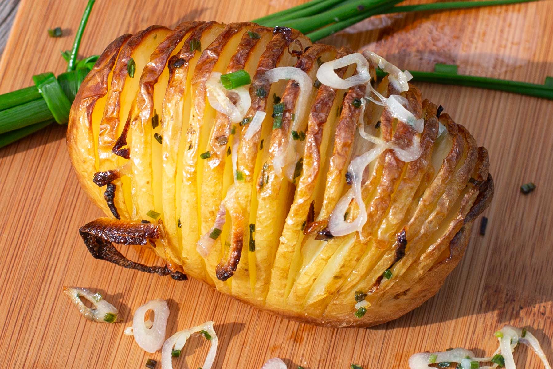 Golden Hasselback potatoes with crispy layers on a wooden board garnished with chives and sliced shallots.