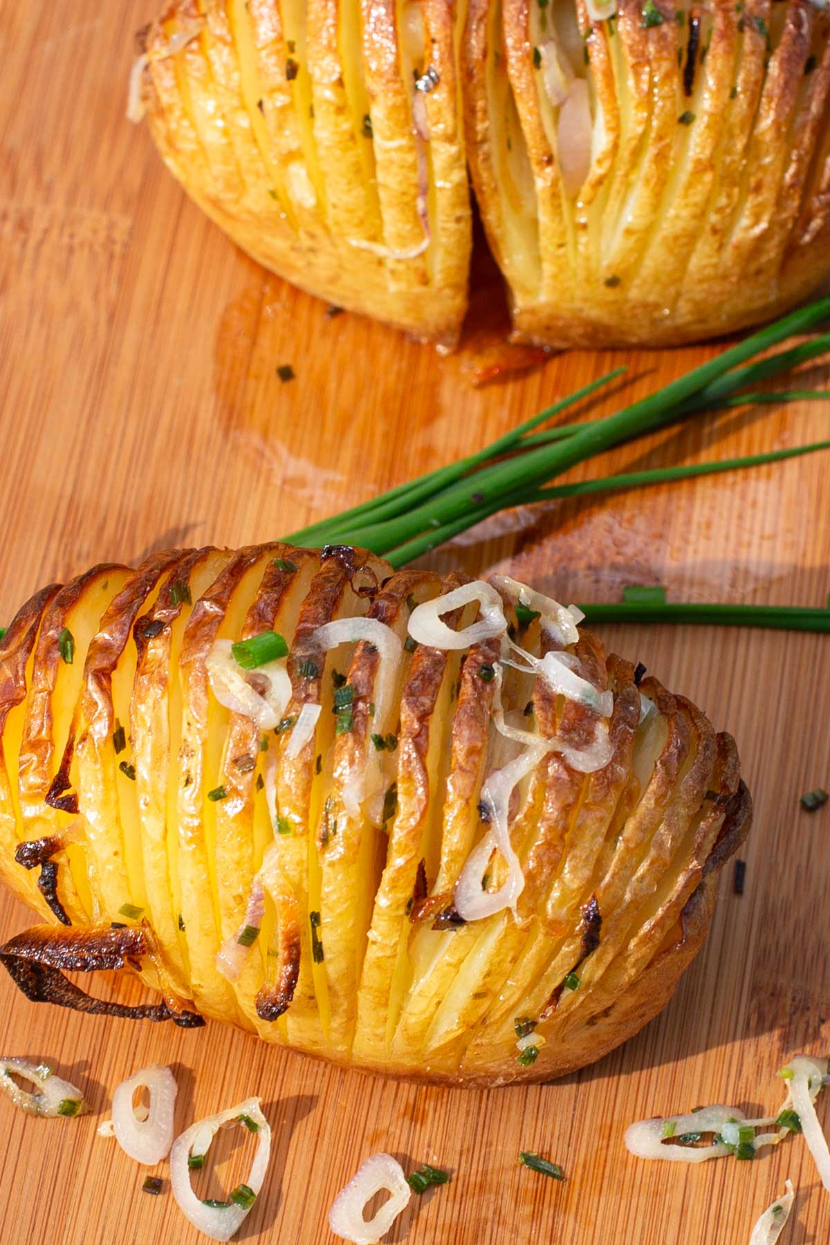 Golden Hasselback potatoes with crispy layers on a wooden board garnished with chives and sliced shallots.