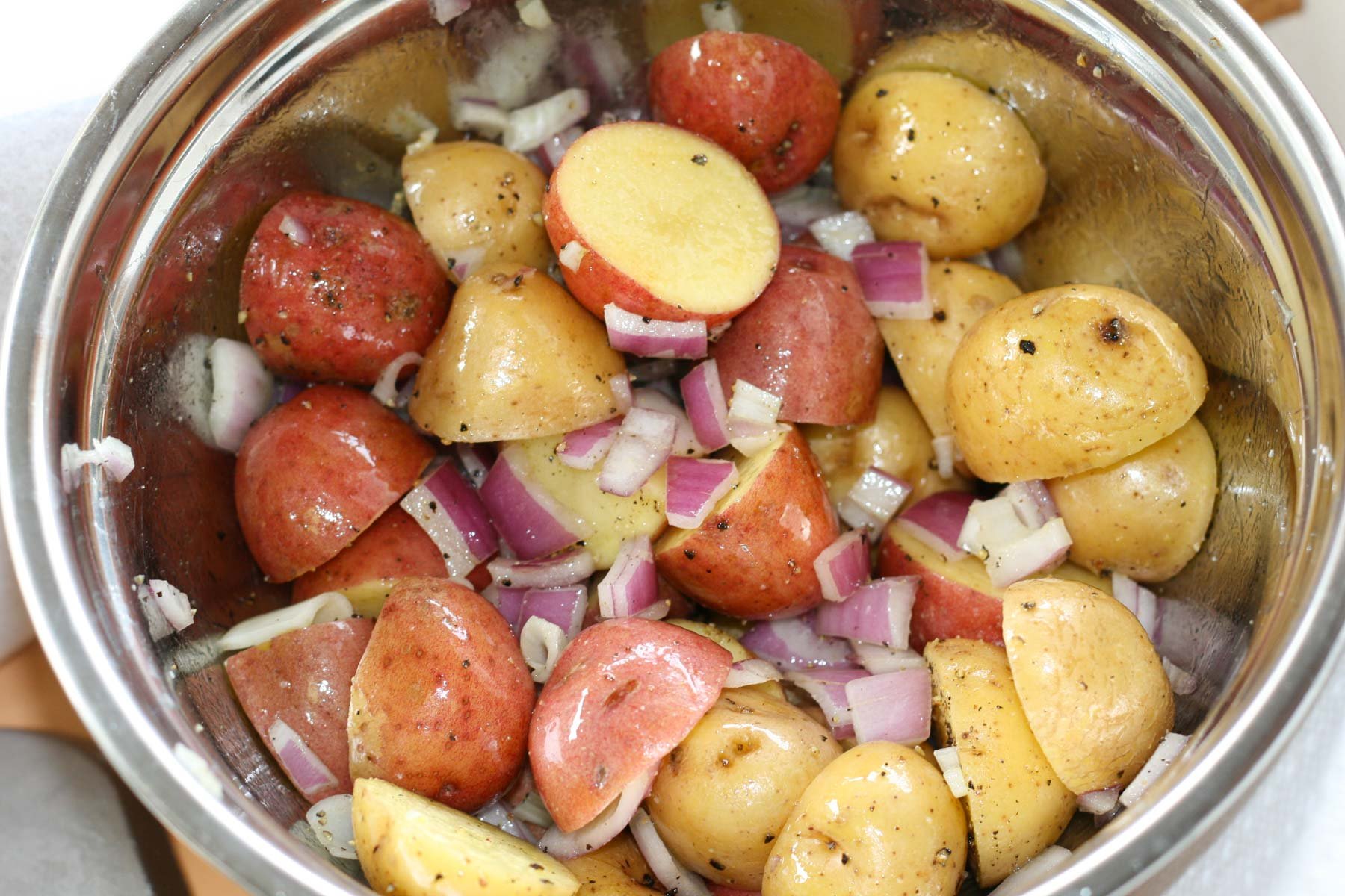 A stainless steel bowl filled with chopped red and yellow potatoes mixed with diced red onions, seasoned with black pepper.