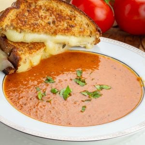Grilled cheese sandwich with melting cheese on golden bread, paired with a bowl of vibrant tomato soup garnished with fresh parsley.