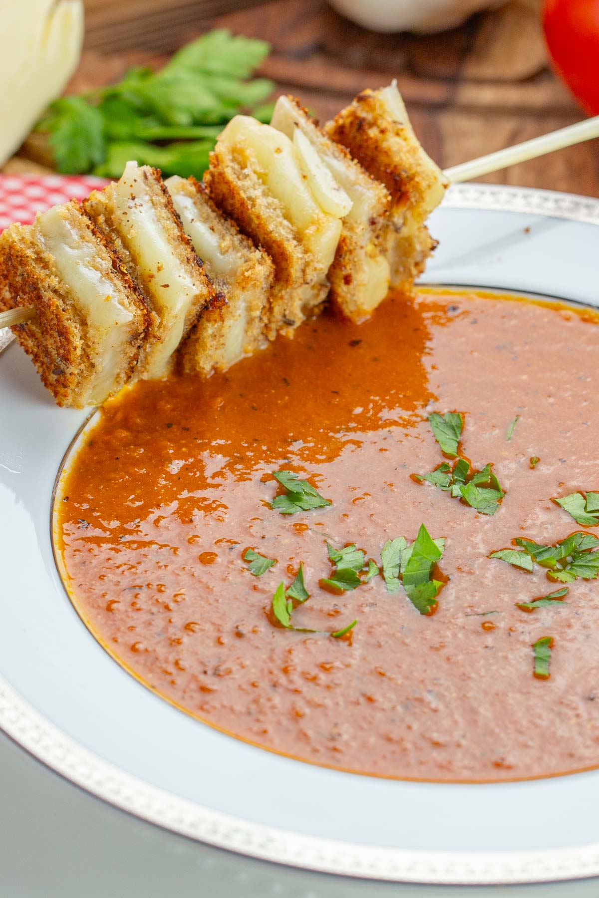 A bowl of vibrant tomato soup garnished with fresh parsley. A skewer of grilled cheese sandwiches rests on the rim.