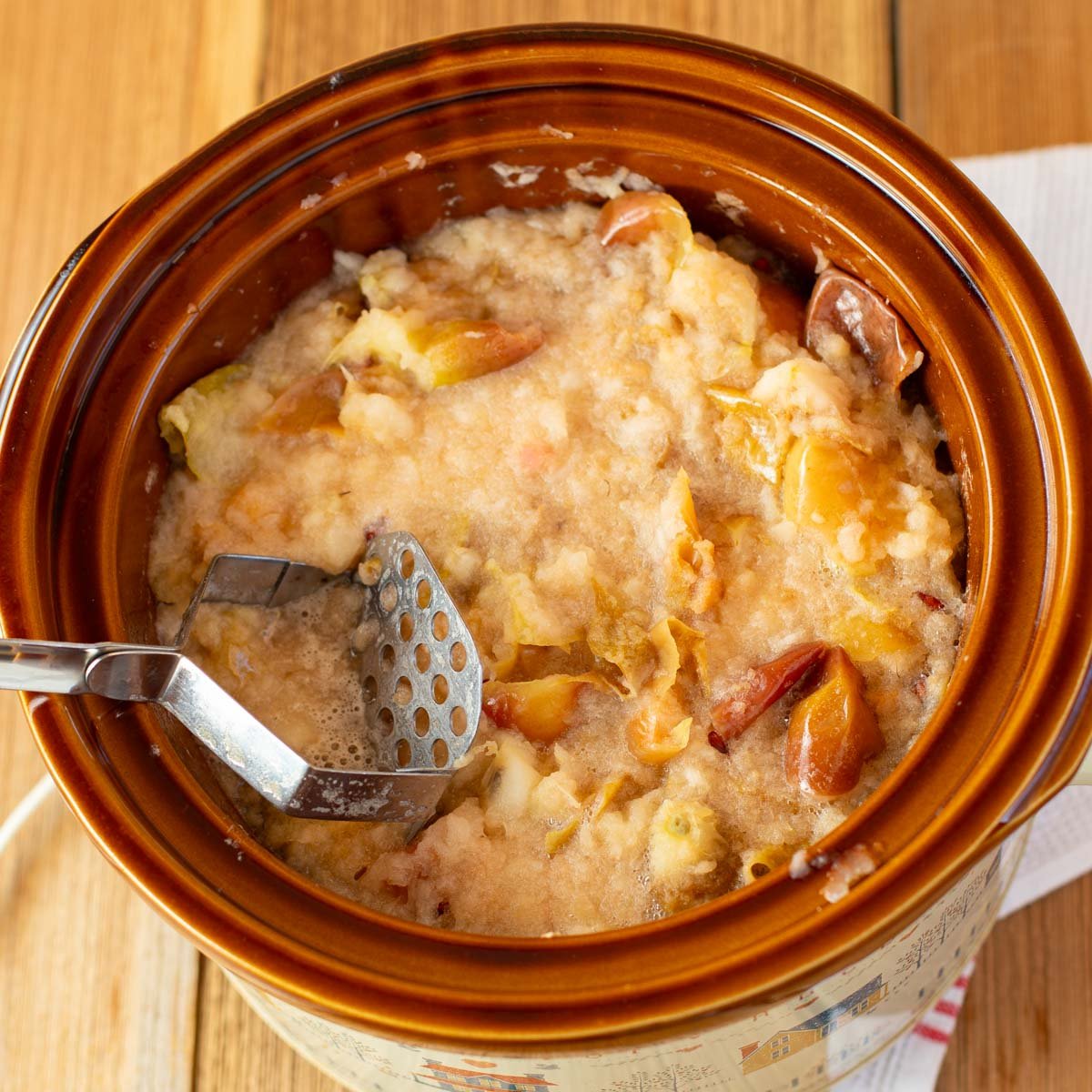 A brown crockpot filled with chunky homemade applesauce. A metal masher is inside, amid soft, partially mashed apple pieces.