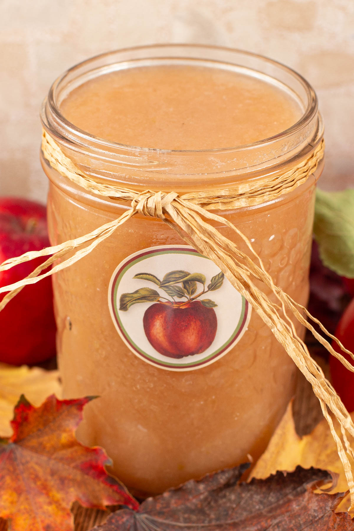 A jar of applesauce wrapped with a raffia bow, featuring an apple label. It's surrounded by red apples and autumn leaves.