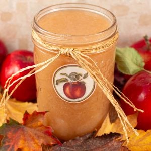 A jar of applesauce wrapped with a raffia bow, featuring an apple label. It's surrounded by red apples and autumn leaves.