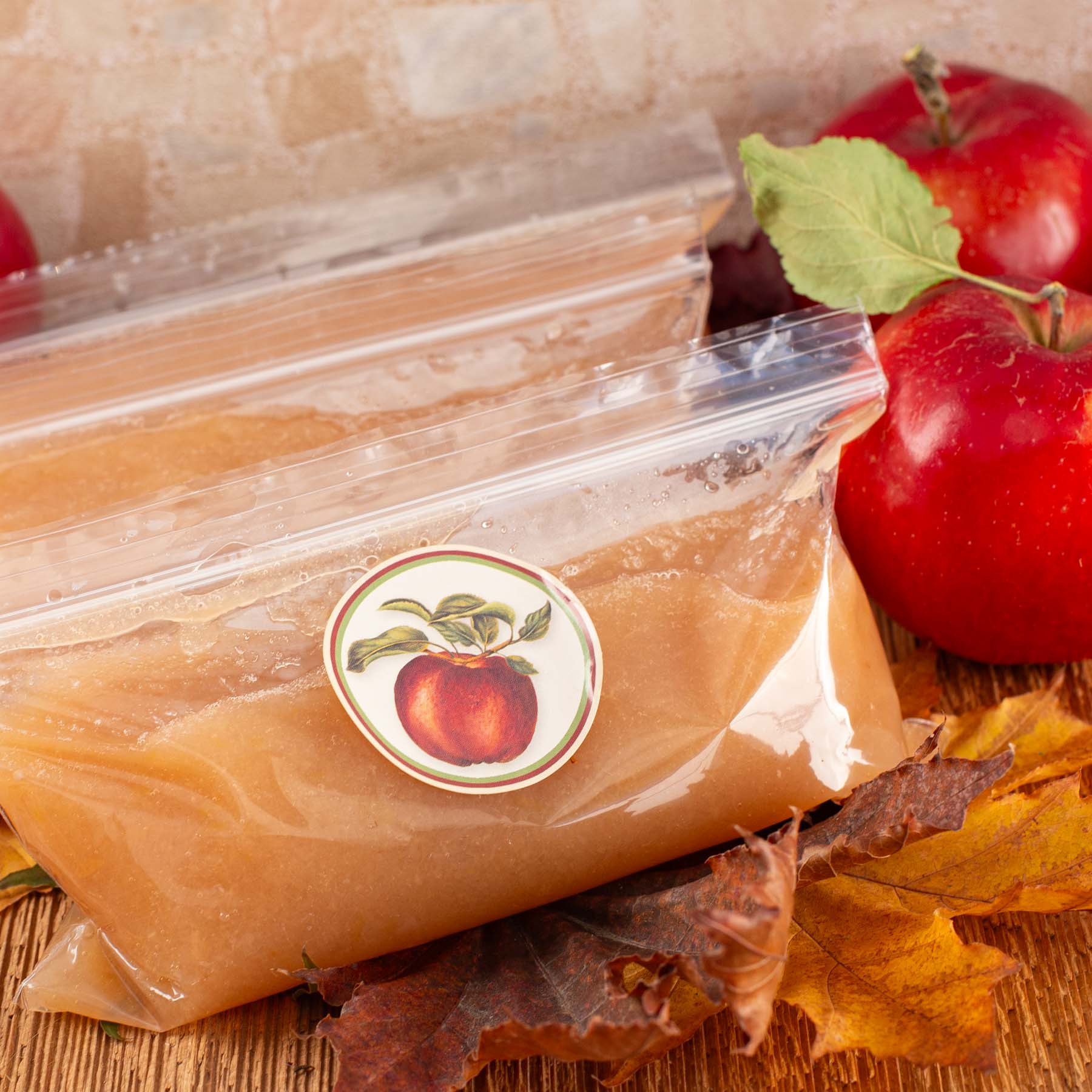 Plastic bags of applesauce with apple stickers rest on wooden surface, surrounded by red apples and autumn leaves.