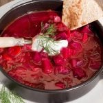 Vibrant red beet soup in a black bowl, topped with dill and a dollop of sour cream, with a slice of bread on the side.