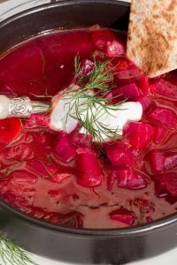 Vibrant red beet soup in a black bowl, topped with dill and a dollop of sour cream, with a slice of bread on the side.