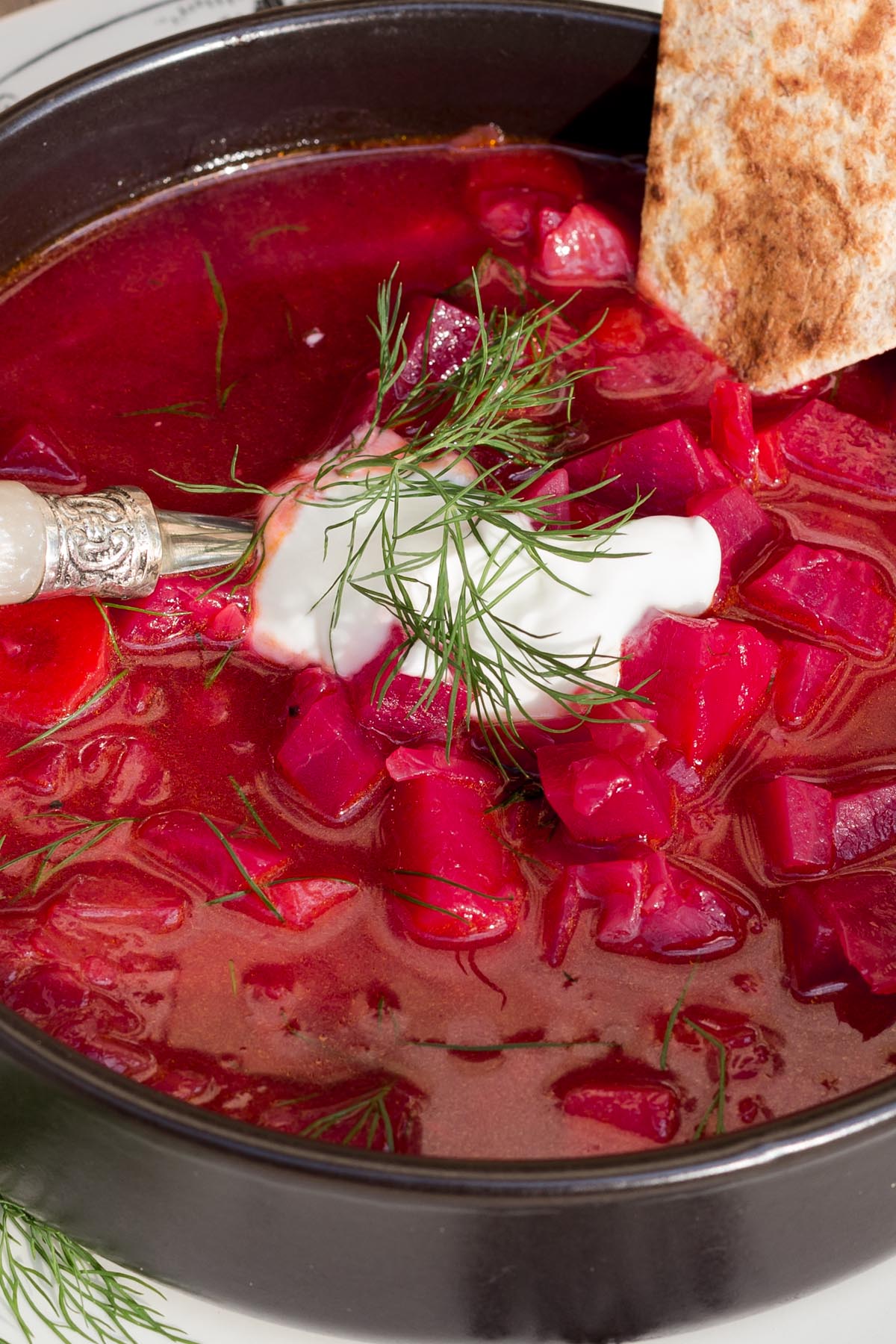 Vibrant red beet soup in a black bowl, topped with dill and a dollop of sour cream, with a slice of bread on the side.