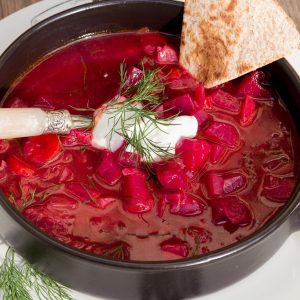 Vibrant red beet soup in a black bowl, topped with dill and a dollop of sour cream, with a slice of bread on the side.