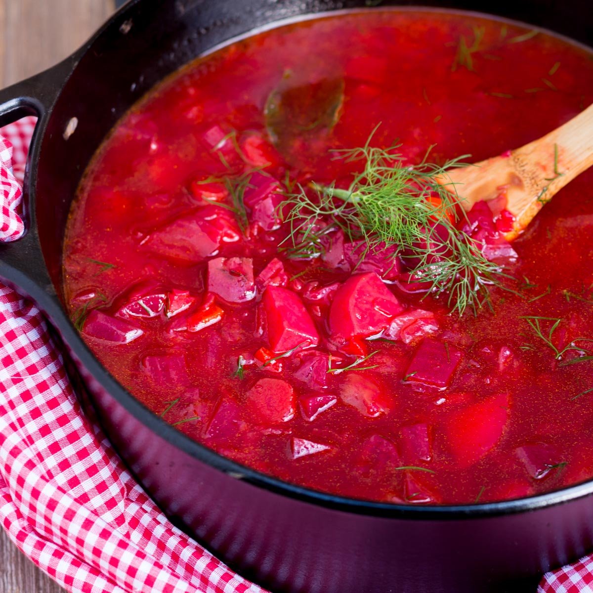Vibrant red beet soup in a black pot, topped with dill, with a slice of bread on the side.