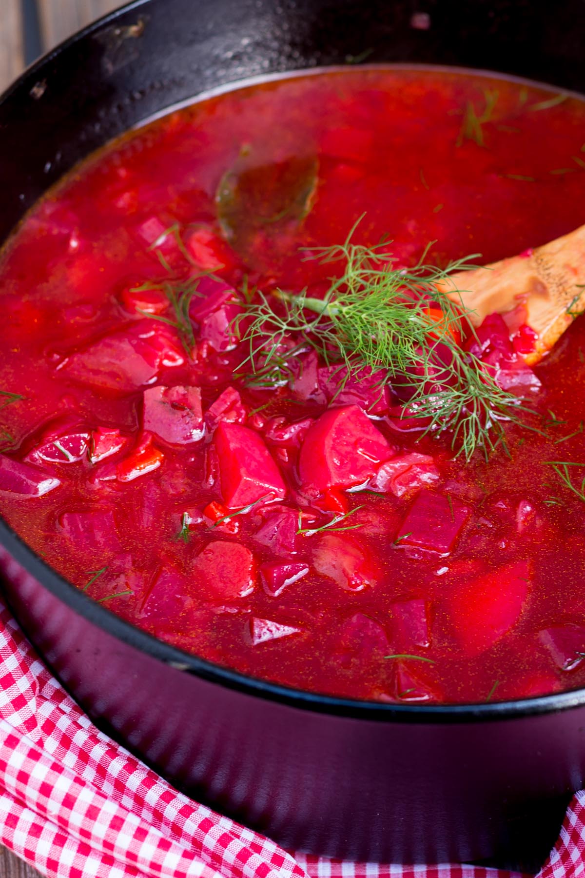 Close-up of vibrant red beet soup in a black pot, topped with dill, with a slice of bread on the side.