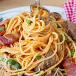 A plate of spaghetti with a fork twirling the pasta, topped with bacon, peas, and mushrooms and garnished with parsley.