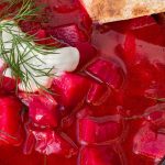 Vibrant red beet soup in a black bowl, topped with dill and a dollop of sour cream, with a slice of bread on the side.