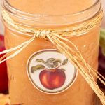 A jar of applesauce wrapped with a raffia bow, featuring an apple label. It's surrounded by red apples and autumn leaves.