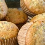Freshly baked zucchini muffins are stacked on a black cooling rack. The muffins have a golden-brown crust, with flecks of green from the zucchini.