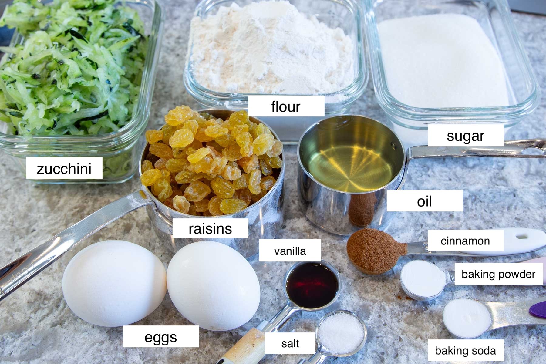 Ingredients for zucchini bread on a counter: grated zucchini, flour, sugar, raisins, eggs, oil, vanilla, cinnamon, salt, baking powder, baking soda.