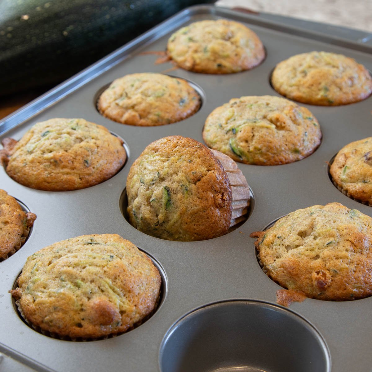 Freshly baked muffins cooling in a metal muffin pan, with one muffin slightly tilted.