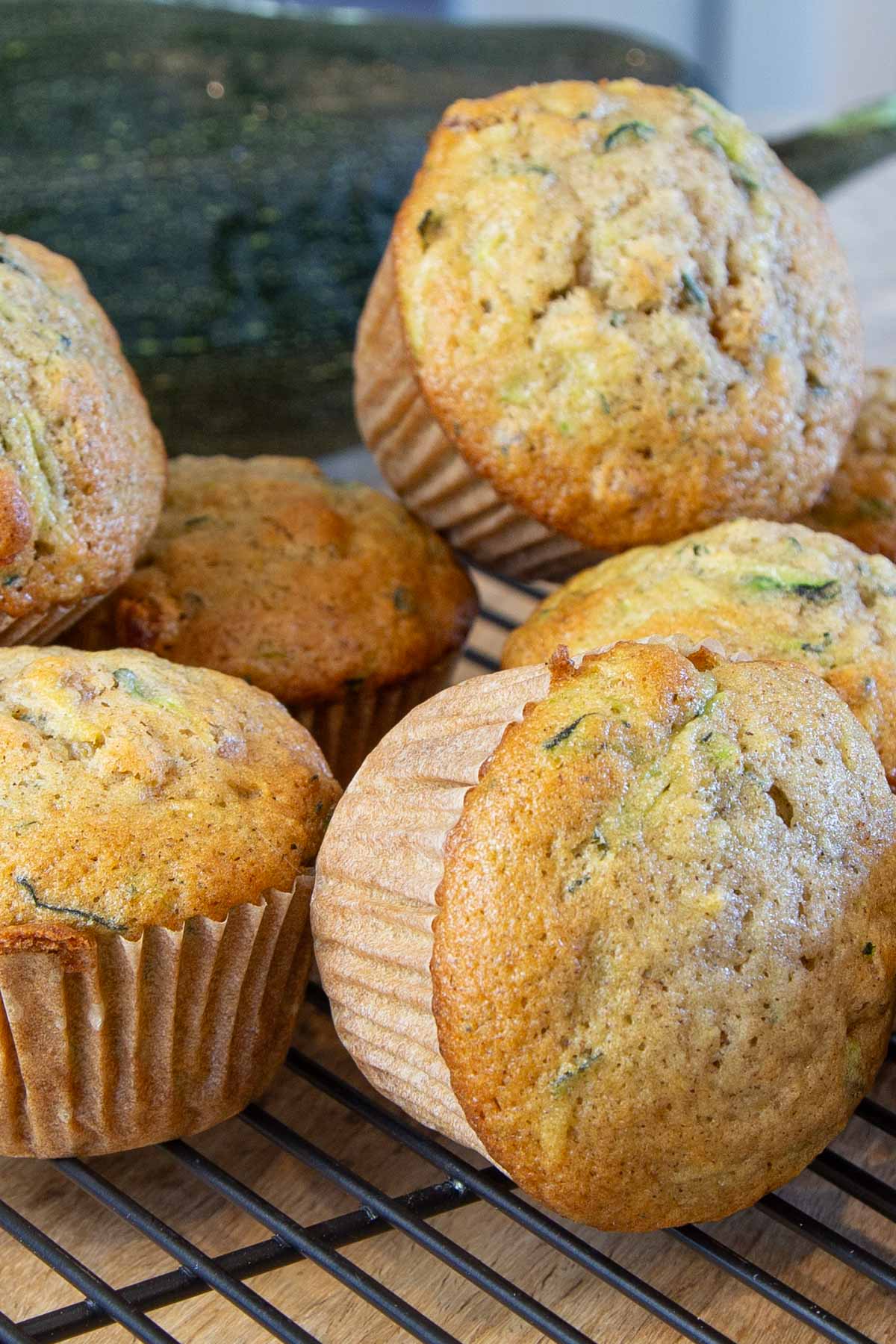 Freshly baked zucchini muffins are stacked on a black cooling rack. The muffins have a golden-brown crust, with flecks of green from the zucchini.
