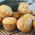 Freshly baked zucchini muffins are stacked on a black cooling rack. The muffins have a golden-brown crust, with flecks of green from the zucchini.