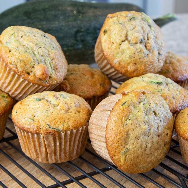 Freshly baked zucchini muffins are stacked on a black cooling rack. The muffins have a golden-brown crust, with flecks of green from the zucchini.