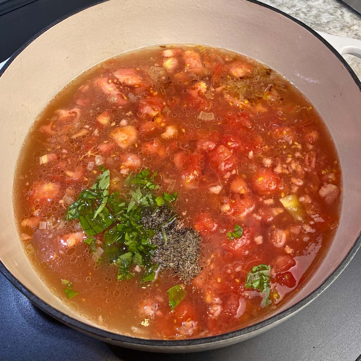 A pot of chunky tomato soup simmers, filled with diced tomatoes, herbs, and spices. Fresh basil leaves and black pepper are visible on the surface.