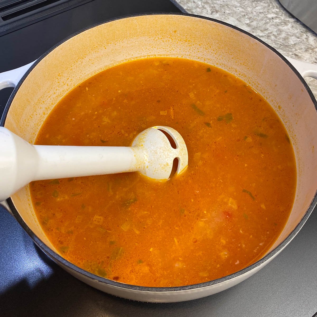 A white immersion blender in a simmering tomato soup within a large pot on a stove.
