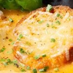 A bowl of tomato soup topped with slices of toasted baguette with melted parmesan, garnished with chopped chives and fresh basil leaves.