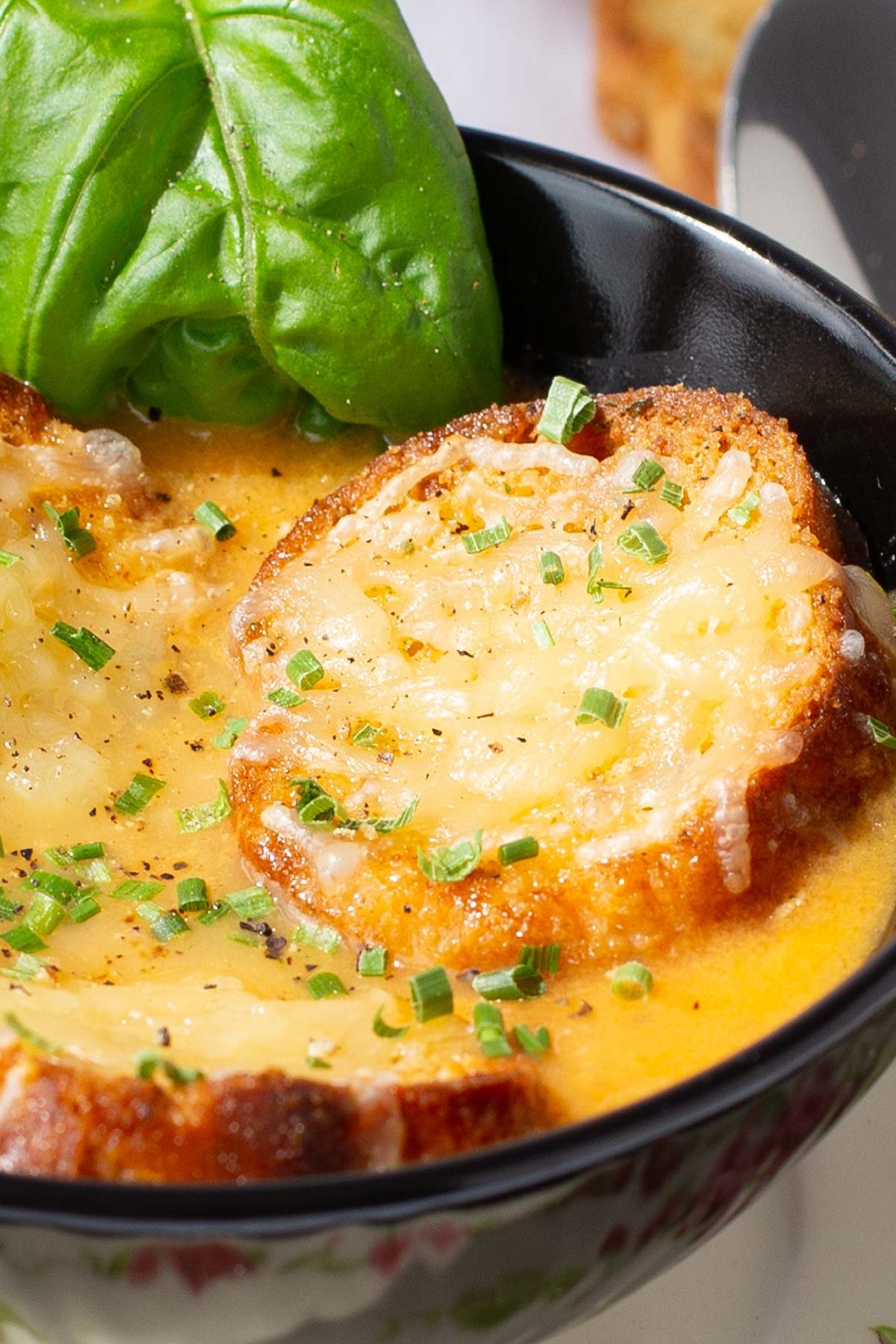 A bowl of tomato soup topped with slices of toasted baguette with melted parmesan, garnished with chopped chives and fresh basil leaves.