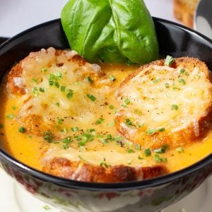 A bowl of tomato soup topped with slices of toasted baguette with melted parmesan, garnished with chopped chives and fresh basil leaves.