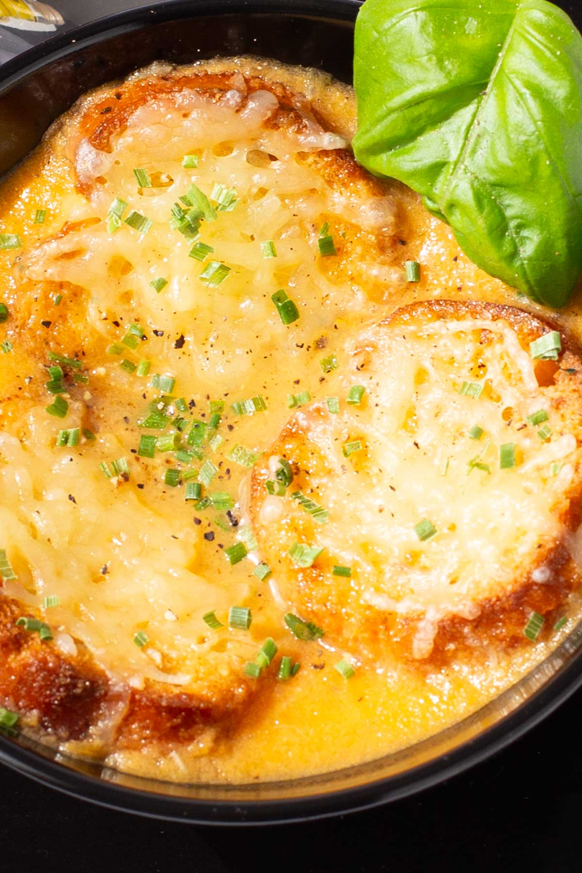 A bowl of tomato soup topped with slices of toasted baguette with melted parmesan, garnished with chopped chives and fresh basil leaves.
